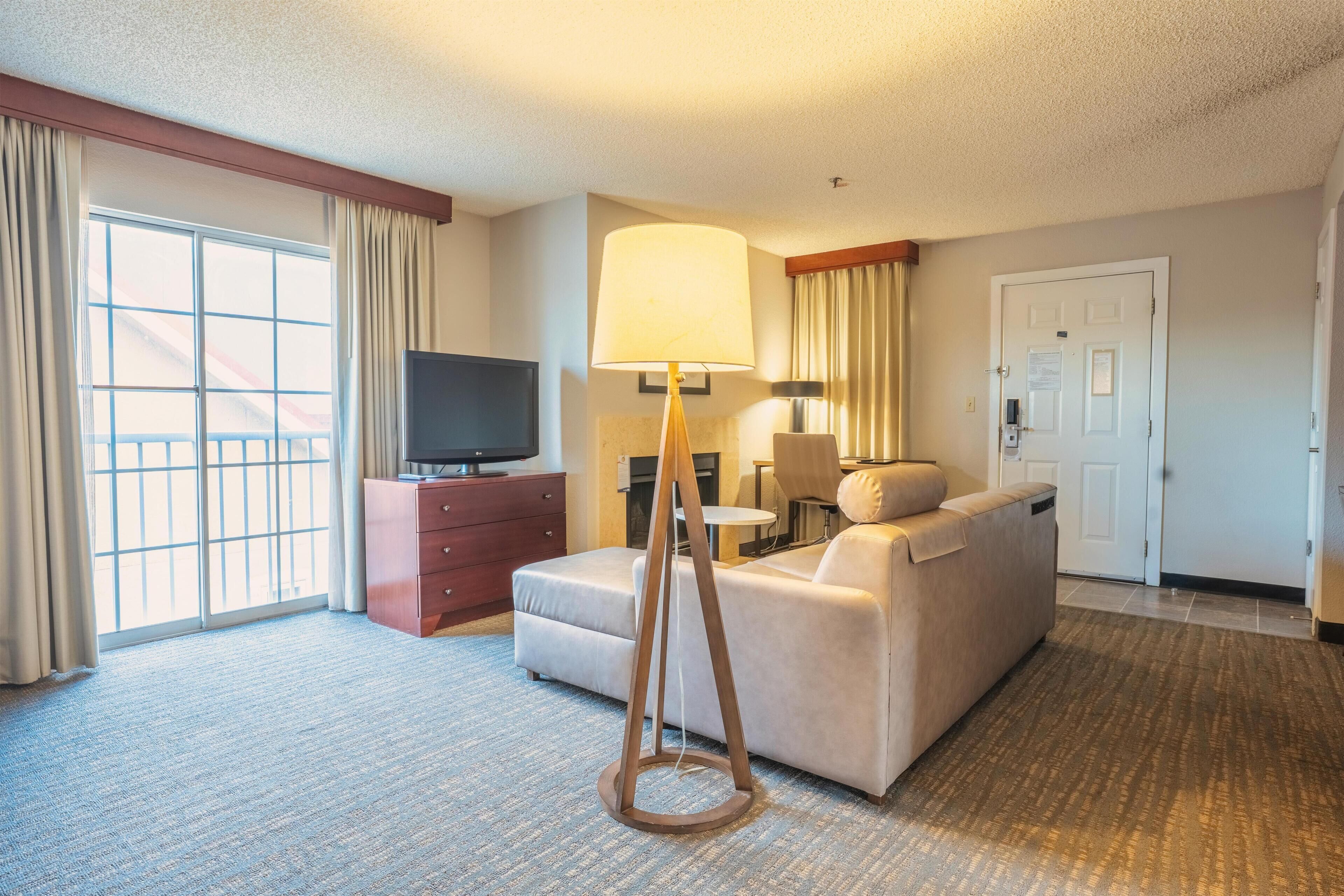 A hotel room with a sofa, a television on a wooden cabinet, a lamp, a door, and a window with curtains.