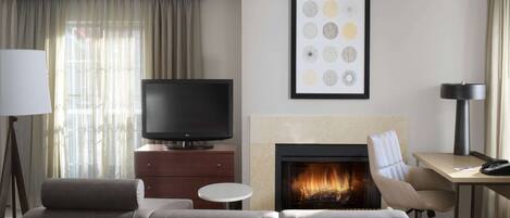 A modern hotel room with a sofa, a flat-screen TV, a fireplace, a desk, and a lamp.