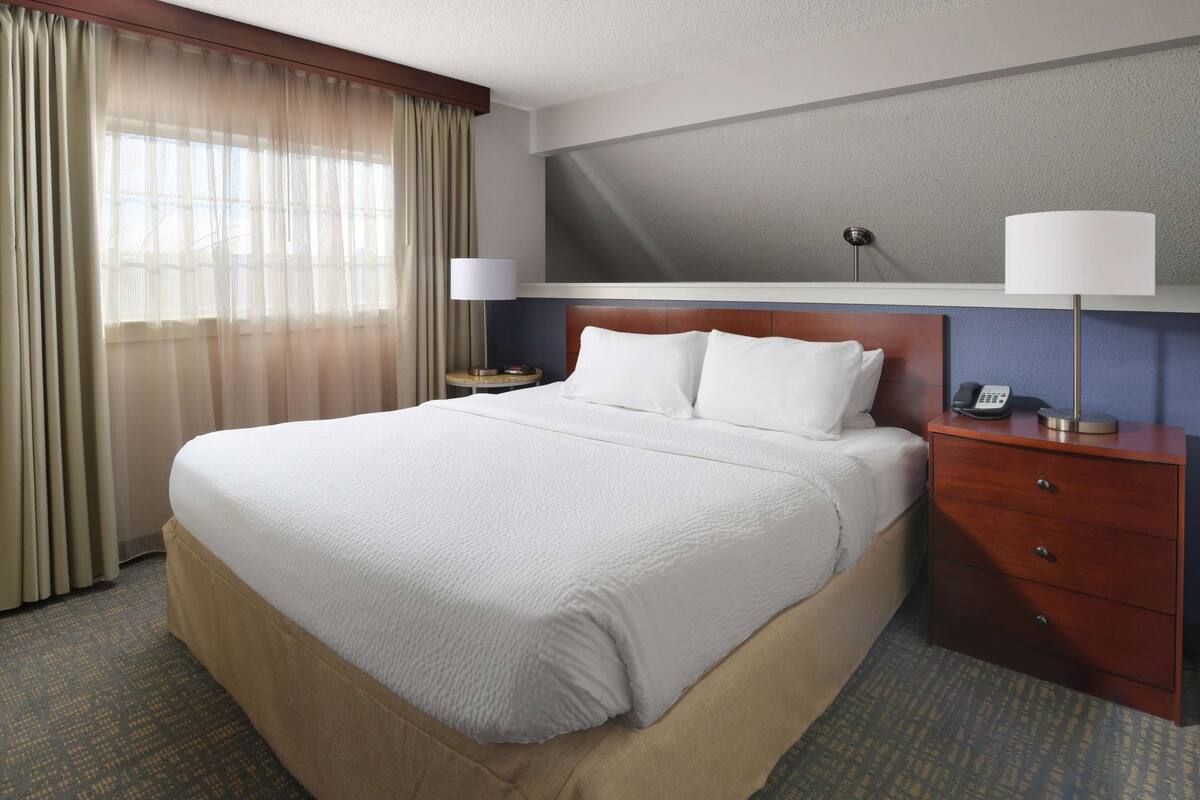 Una habitación de hotel con una cama grande, una mesita de noche con lámpara, teléfono y una ventana con cortinas.