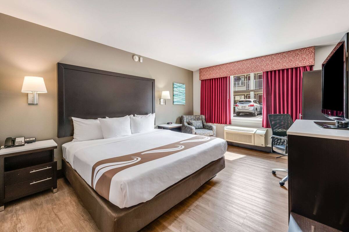 A hotel room with a large bed, a desk, a chair, a lamp, and a window with curtains.