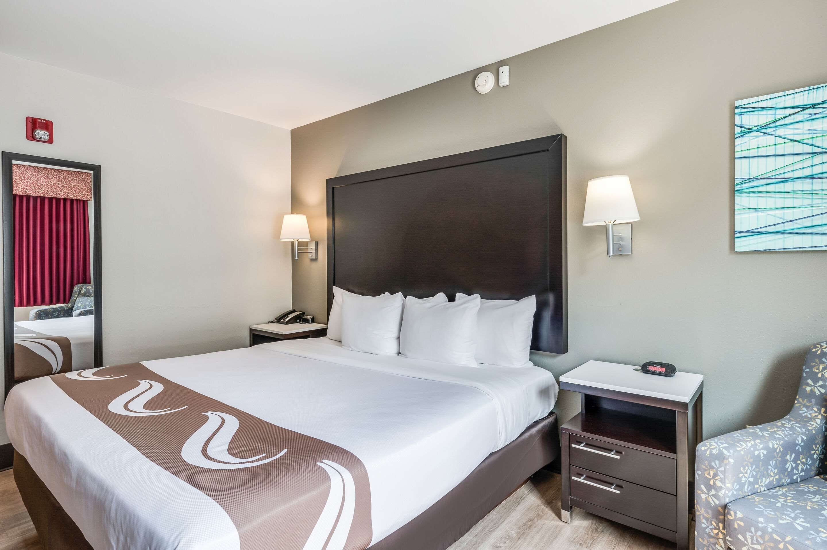 Habitación de hotel con una cama grande, una mesita de noche con lámpara, una silla y un cuadro en la pared.