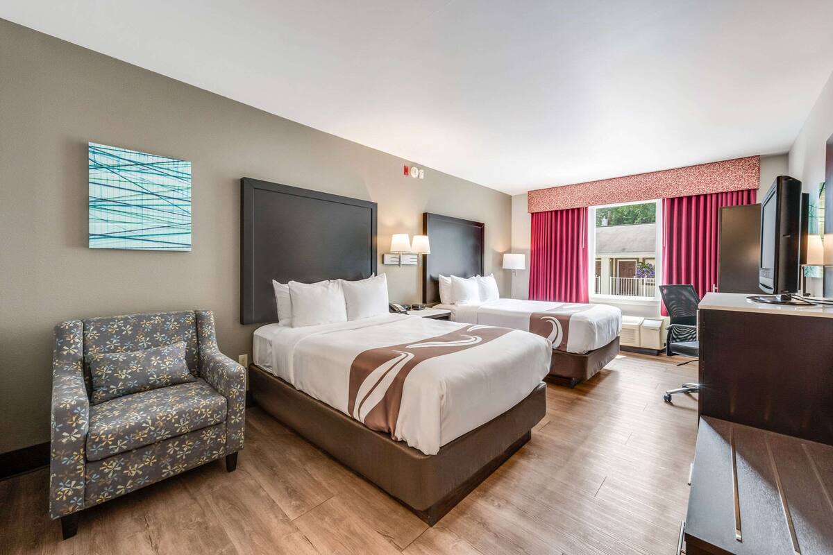 A hotel room with two beds, a desk, a chair, and a TV.