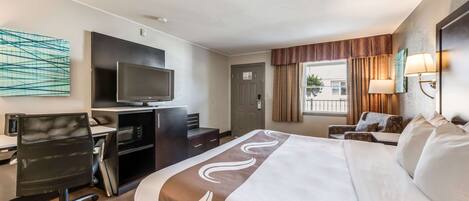A hotel room with a large bed, a desk with a computer, a TV, and a window with curtains.