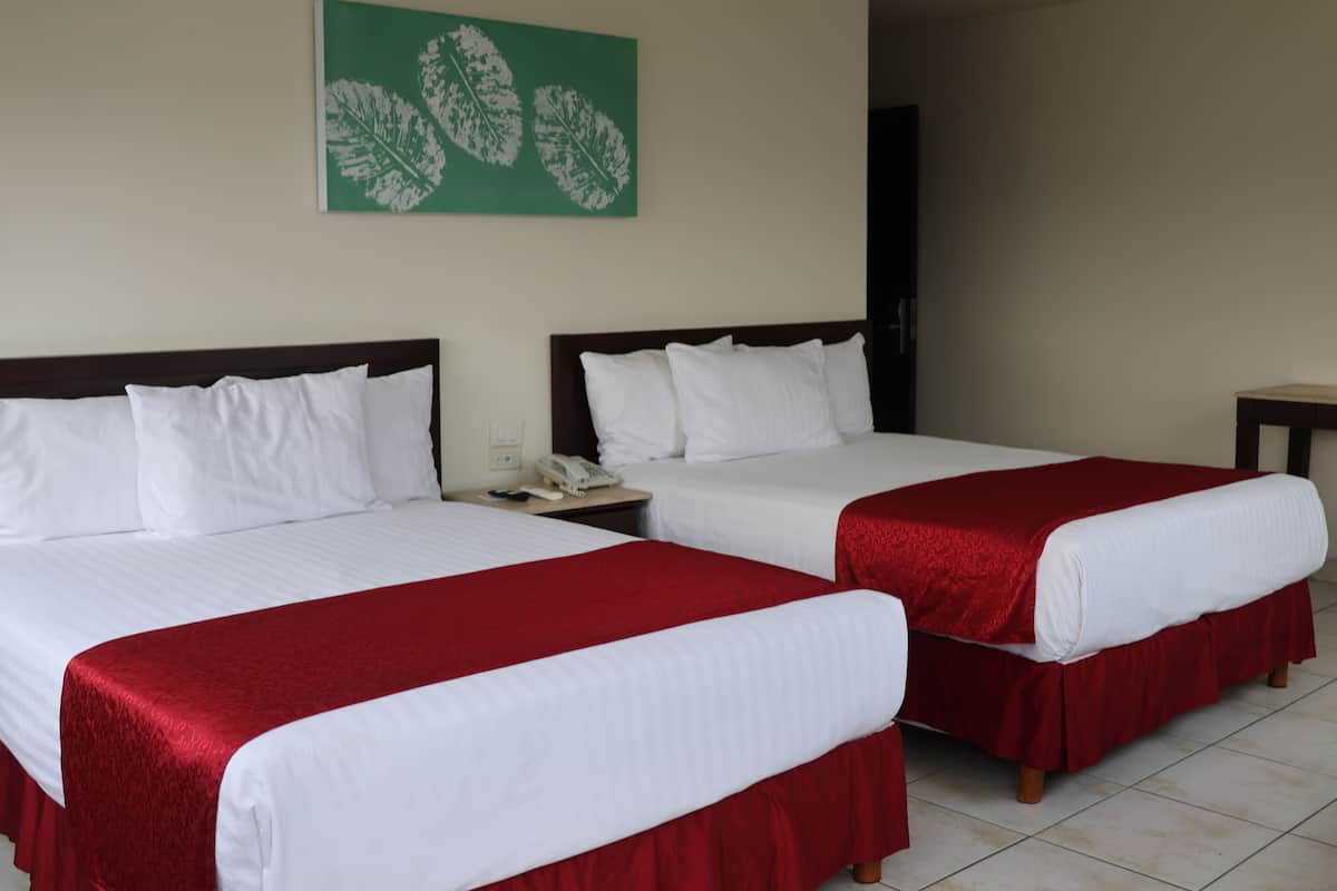 A hotel room with two beds, each with white linens and red bedspreads. There is a green artwork on the wall and a small table with a phone and water bottles.