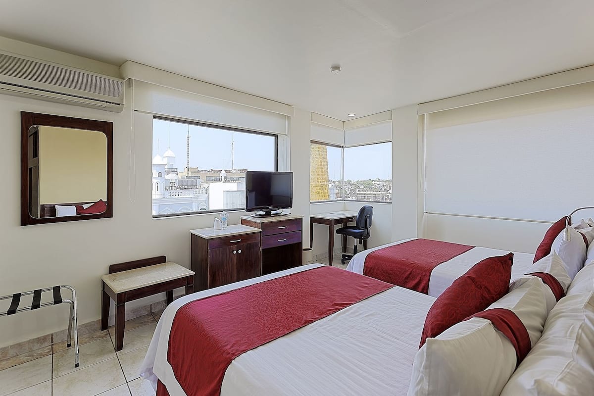 A hotel room with two beds, a desk, a chair, a TV, and a window with a city view.