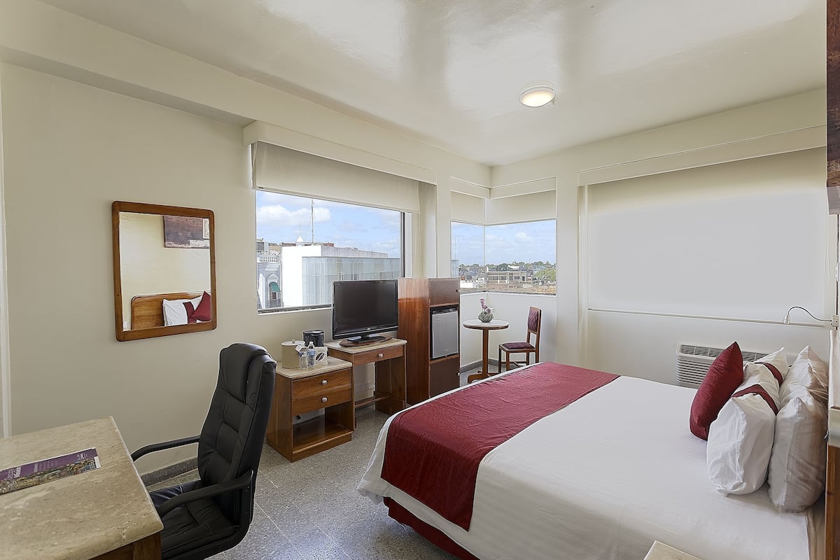A hotel room with a bed, desk, chair, television, and windows with a view.