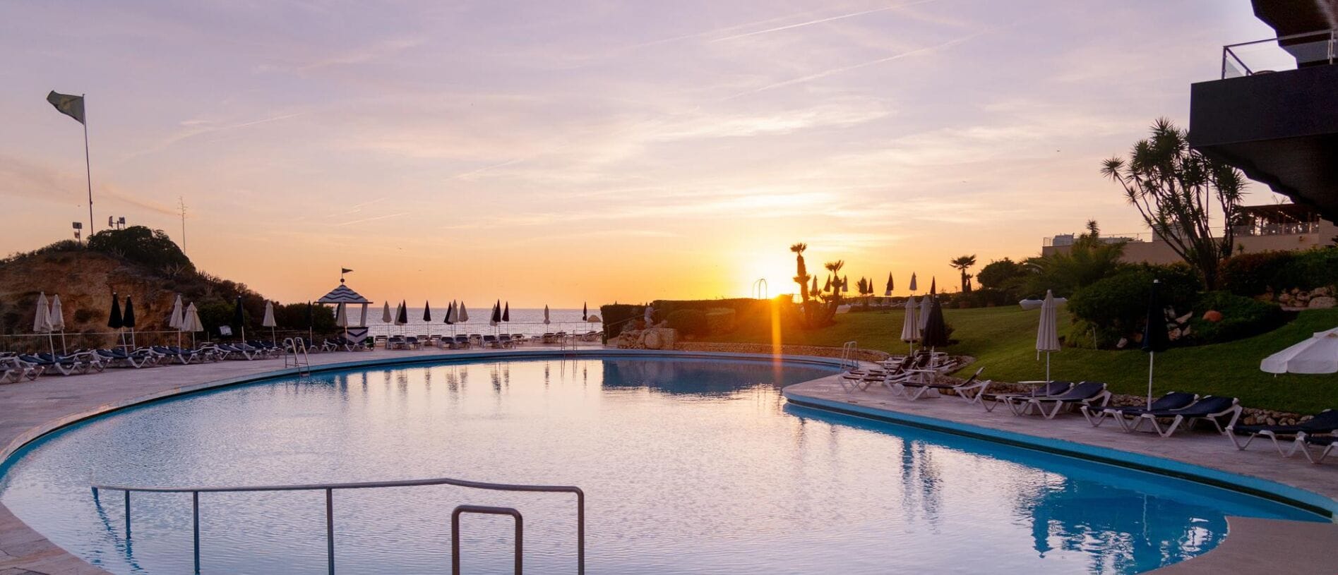 Una piscina con sillas de descanso y sombrillas, rodeada de follaje y un atardecer al fondo.
