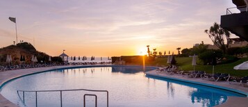 A swimming pool with lounge chairs and umbrellas, surrounded by greenery and a sunset in the background.