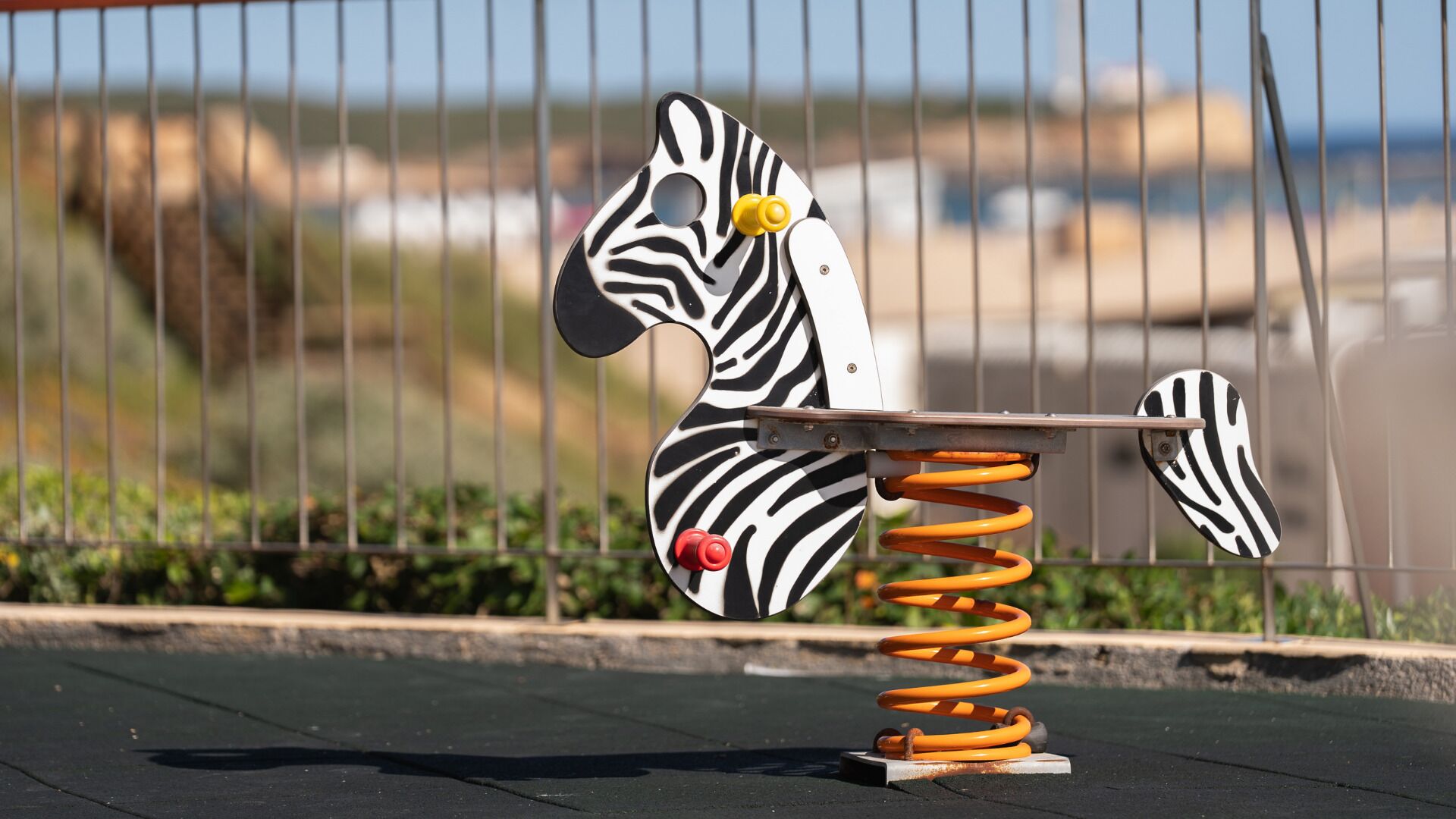 A zebra-shaped swing with a metal frame and orange springs, set against a blurred background of a fence and greenery.
