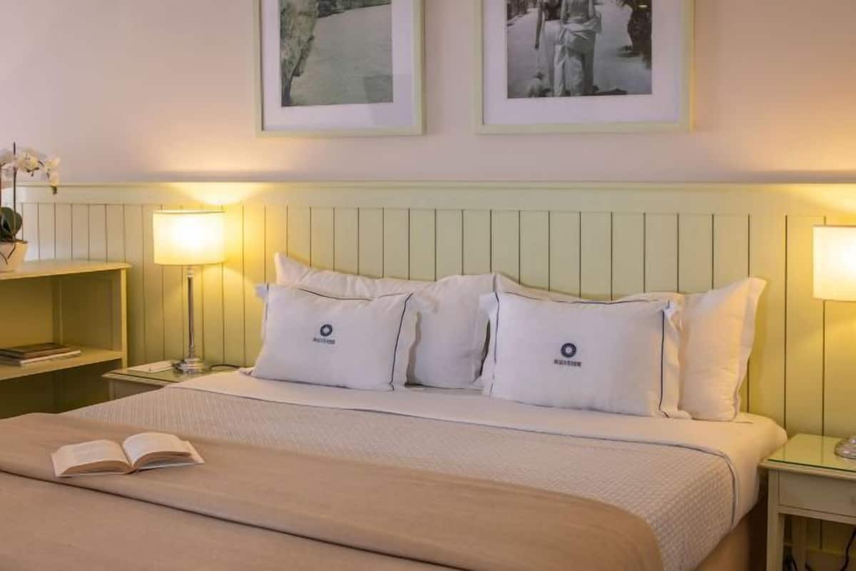 Een hotelkamer met een bed, twee bedlampjes, een nachtkastje met een lamp, een lijst aan de muur en een boek op het bed.