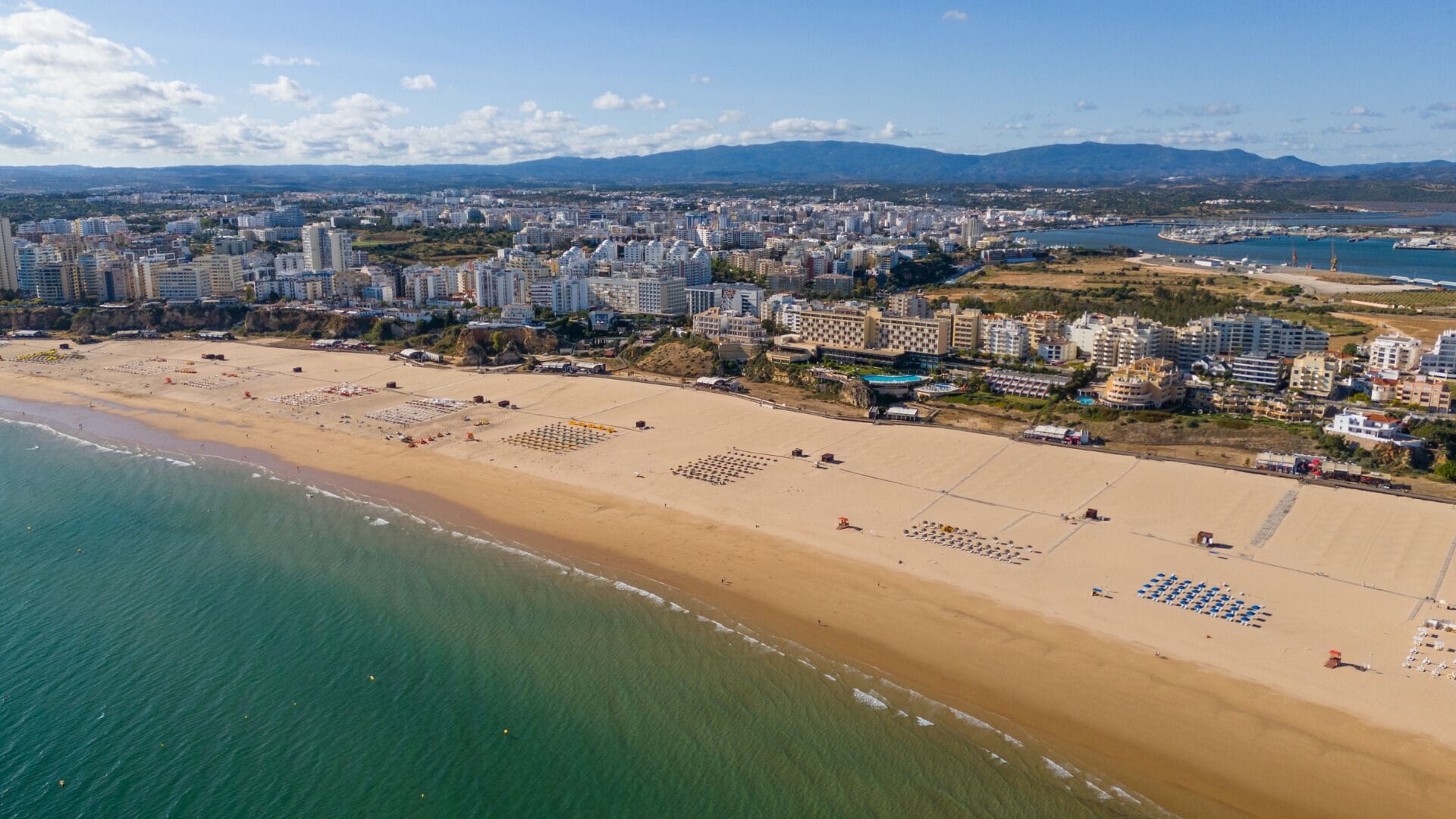 An aerial view of a coastal city with a wide sandy beach, numerous high-rise buildings, and a marina.