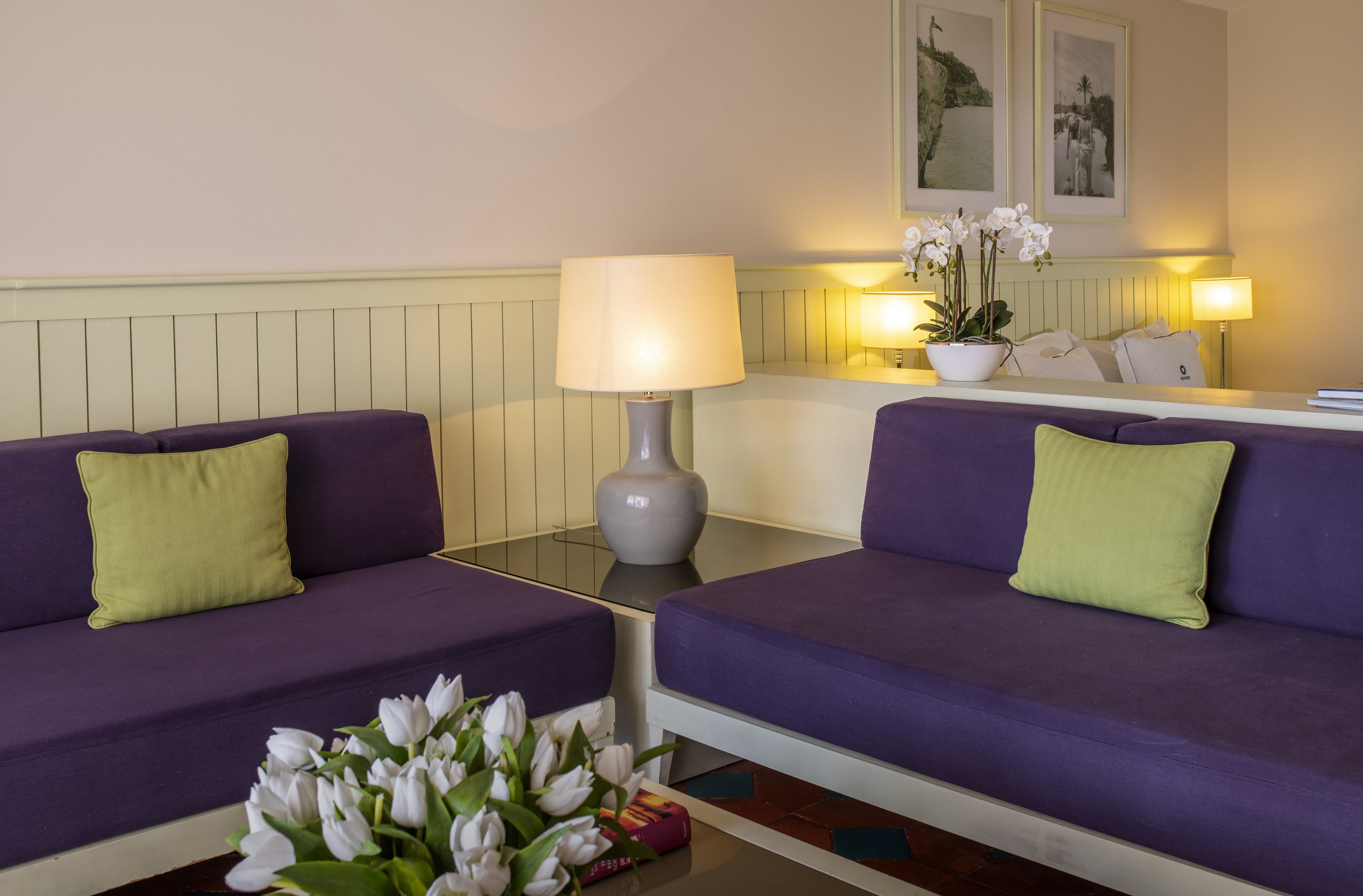 A room with two purple sofas, a small table with a lamp, and a vase with white flowers.