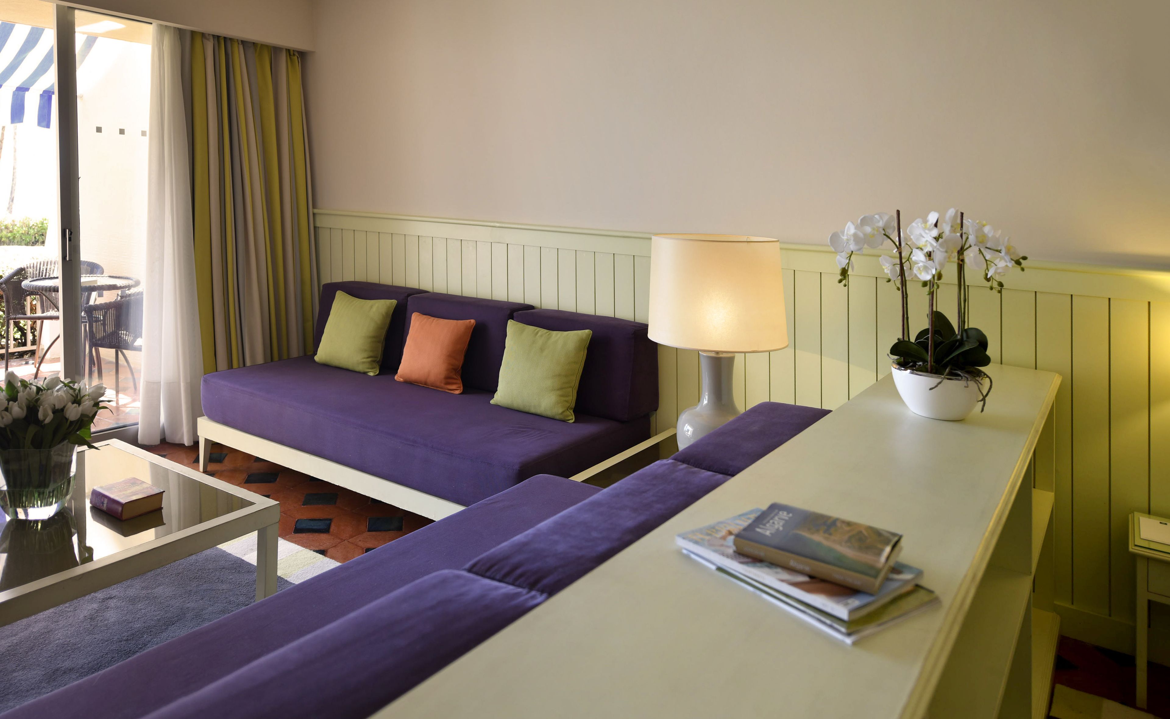 Quarto com um sofá roxo, uma mesa lateral branca, um abajur e um vaso com planta.