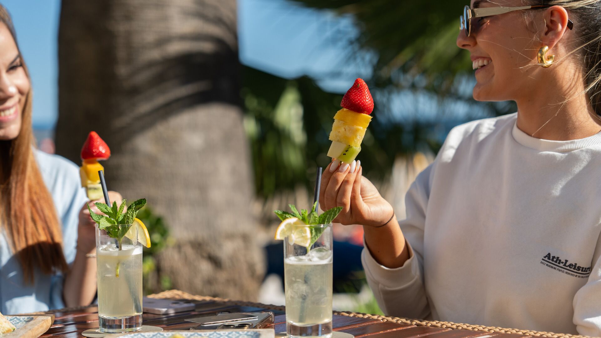 A person holding a dessert with a strawberry on top, a drink with a mint garnish, and another drink with a lemon slice, all on a table with a palm tree in the background.