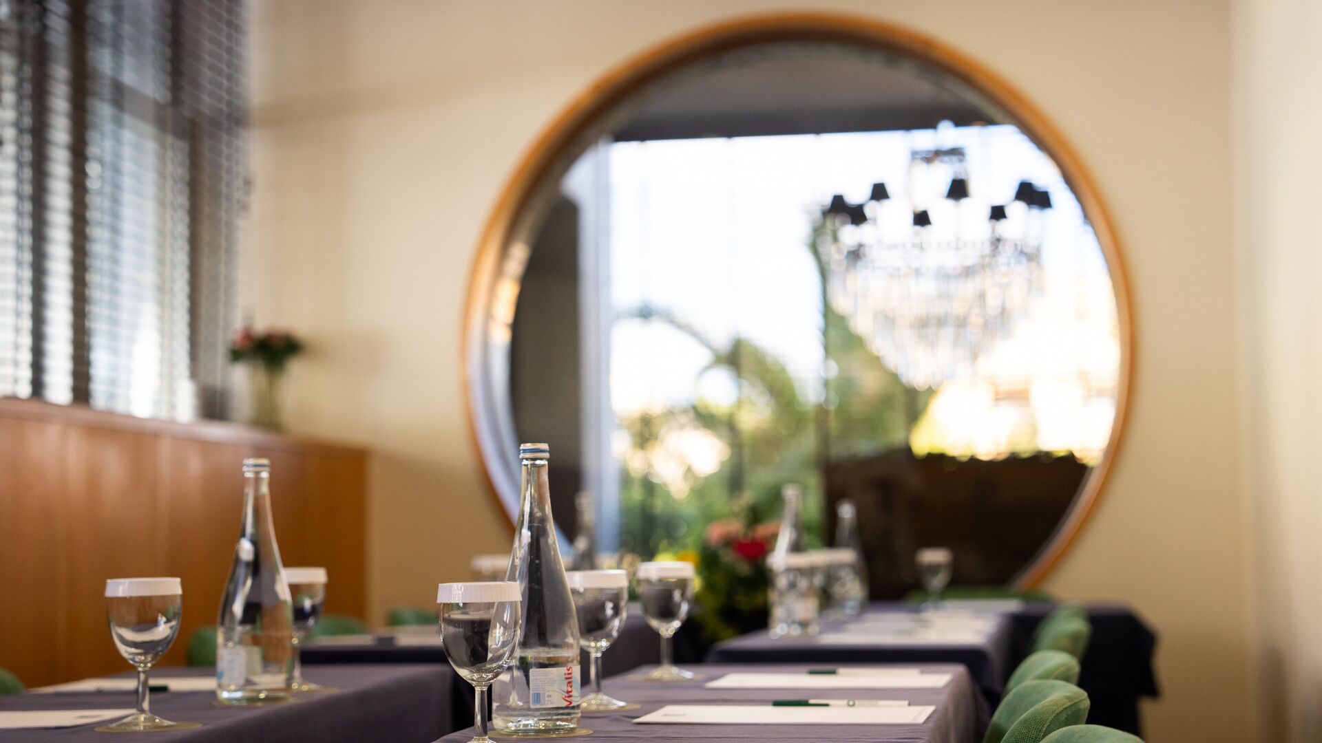 Une table à manger avec des verres et des bouteilles de vin, un miroir qui reflète un lustre, et une vue sur la verdure à l’extérieur.