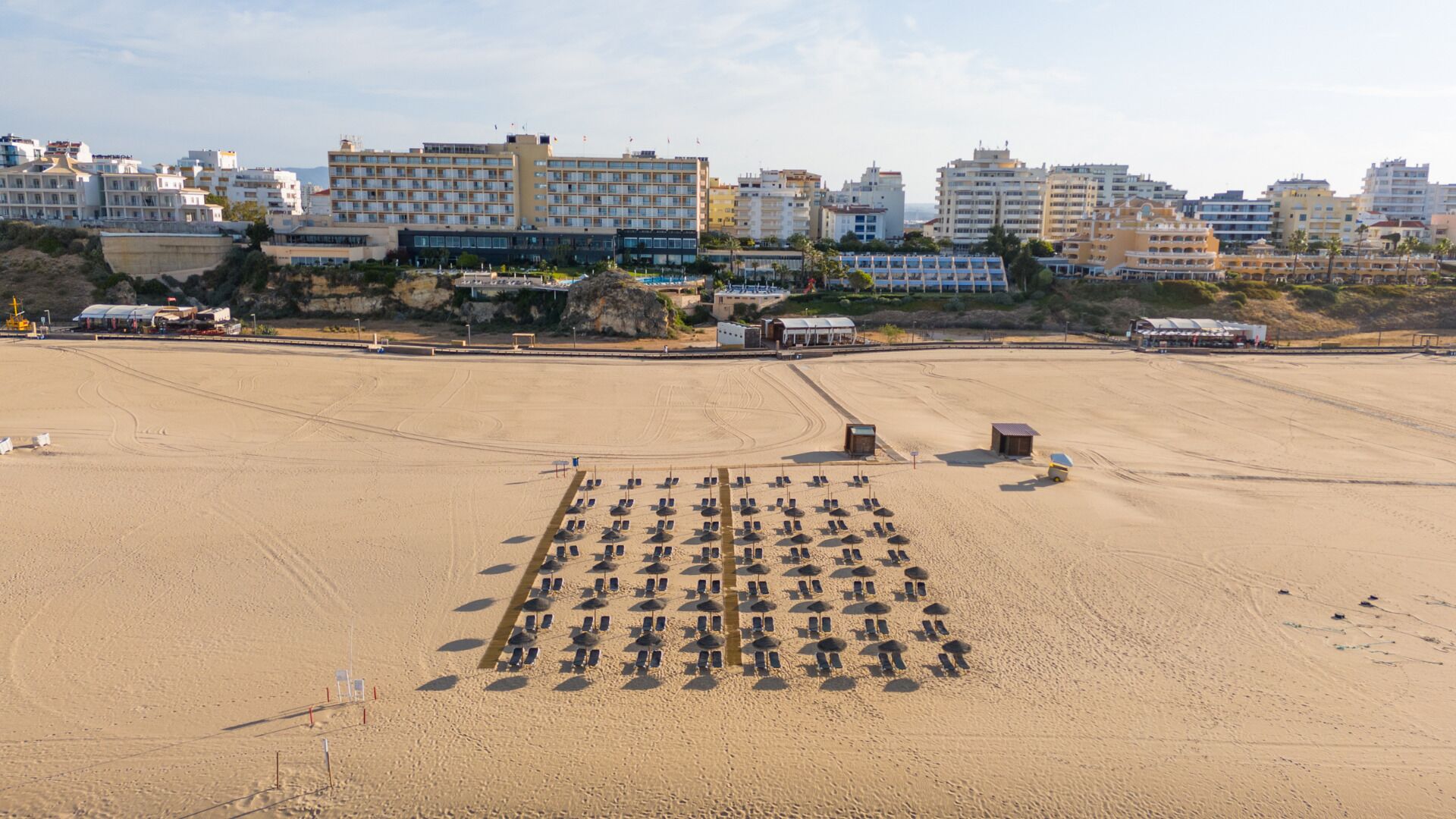Ein Strand mit Reihen von Liegestühlen und Sonnenschirmen, Gebäuden im Hintergrund und einer Sandfläche mit vereinzelten Bauten.