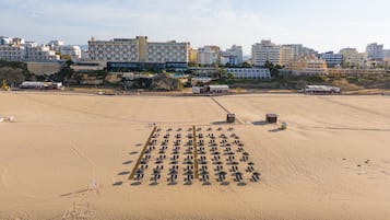 Uma praia com fileiras de espreguiçadeiras e guarda-sóis, edifícios ao fundo e uma área arenosa com algumas construções.