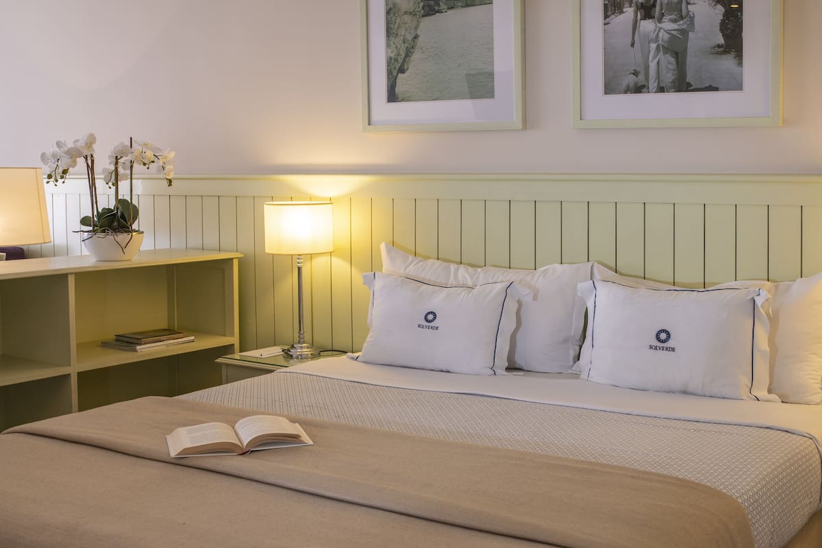 Een keurig opgemaakt hotelkamer met een bed, bedlampjes, een nachtkastje, een boekenplank en een lijst aan de muur.