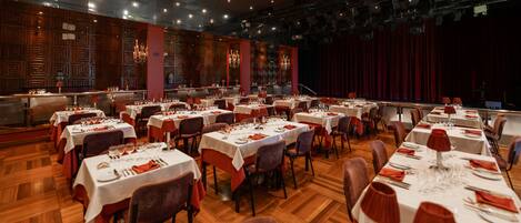 Un amplio salón de banquetes con varias mesas redondas dispuestas para comer, con manteles blancos y servilletas rojas.