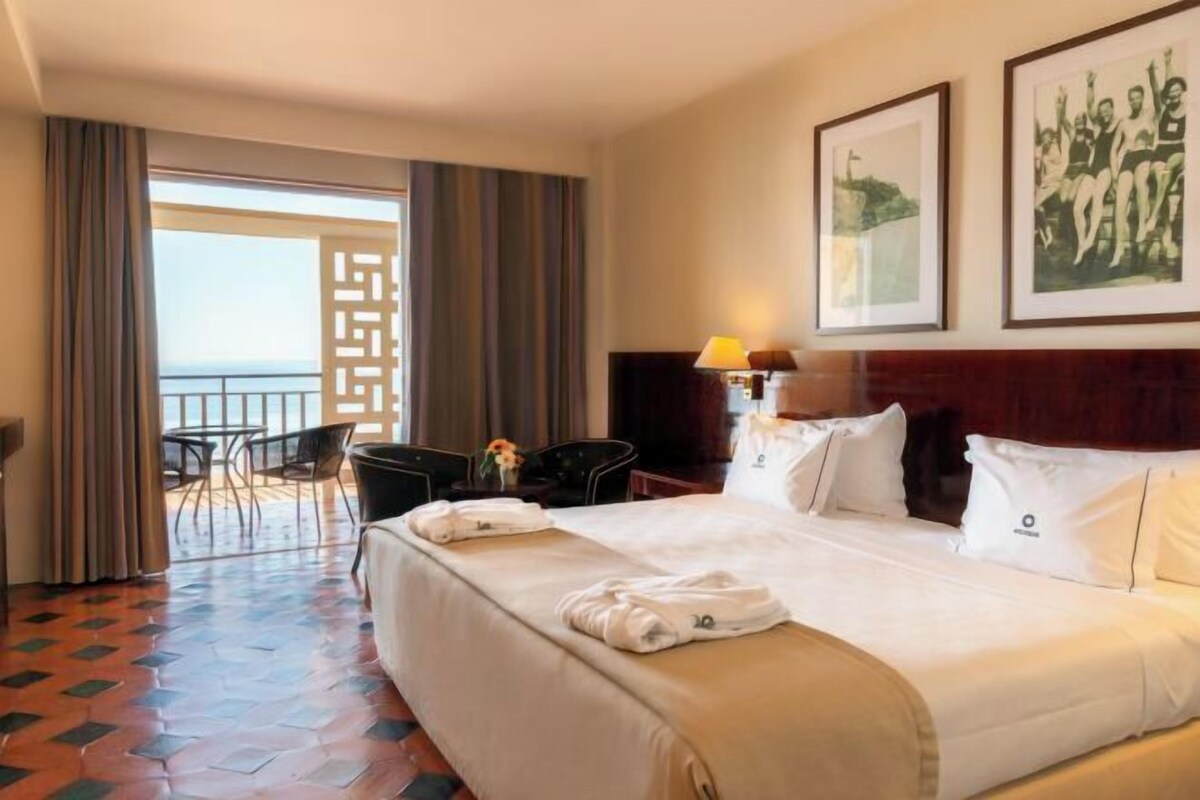 A hotel room with a large bed, a desk, a chair, a balcony view, and two framed pictures on the wall.