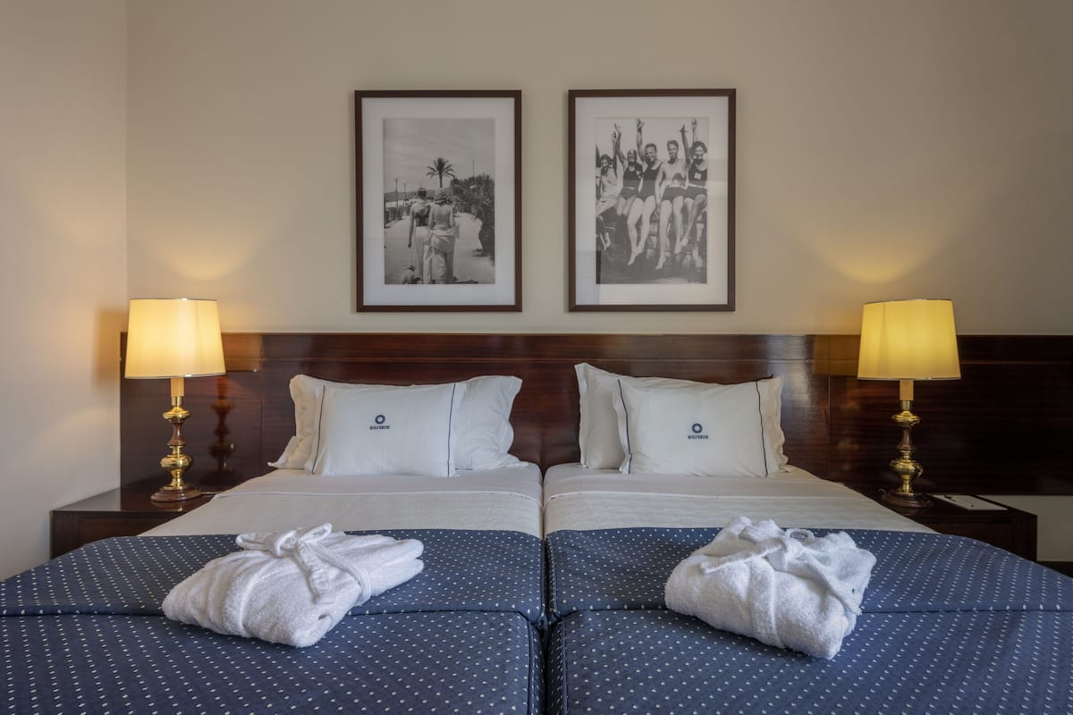Een tweepersoonsbed met twee kussens, een hoofdbord, twee nachtkastjes met lampen, en twee foto's in lijsten aan de muur.