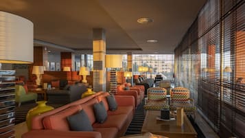 A modern hotel lobby with a long sofa, armchairs, and a coffee table. There are lamps and a large window with blinds.