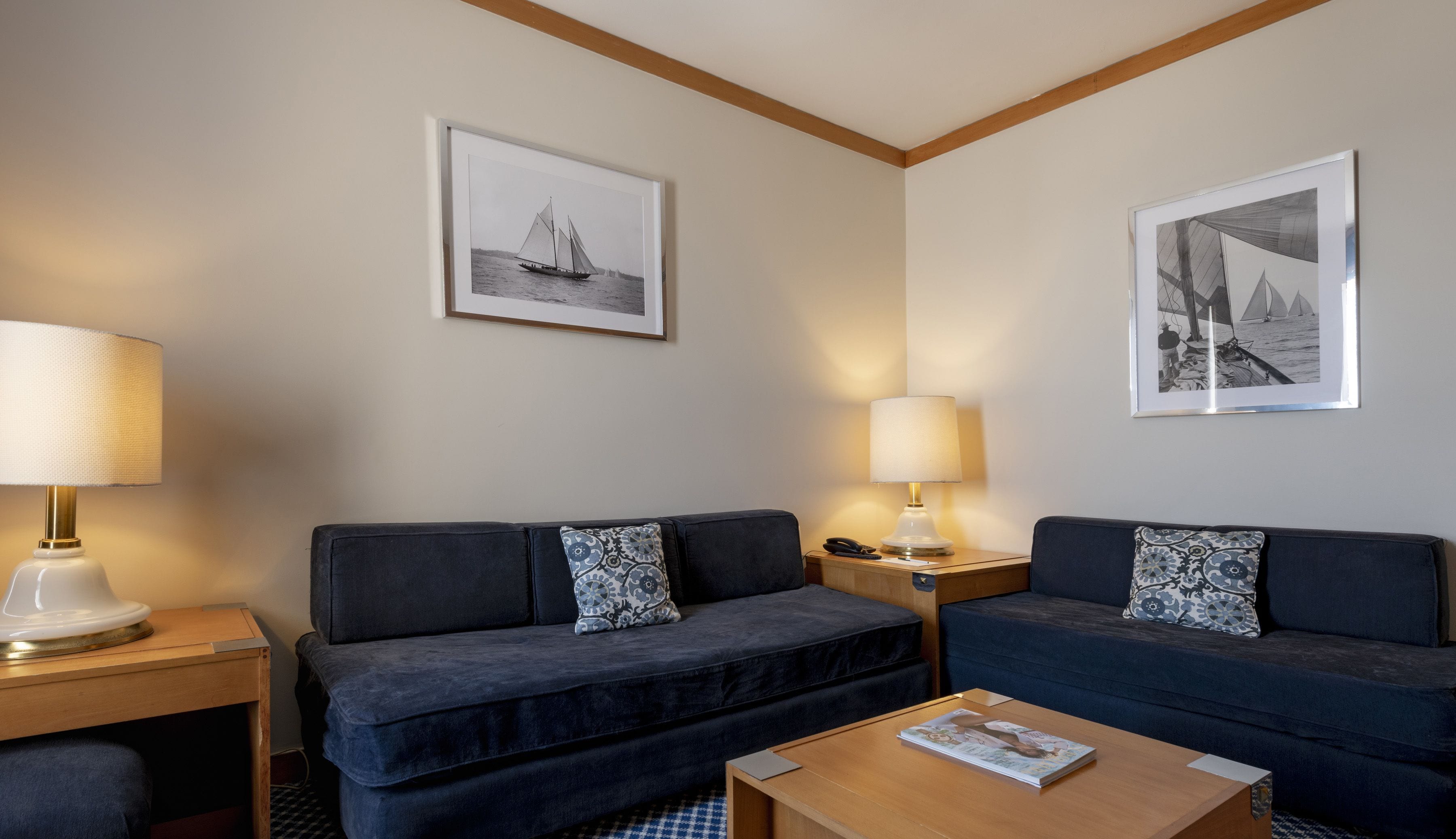A hotel room with two dark blue sofas, a wooden coffee table, and two bedside lamps. There are two framed black and white photographs on the walls.