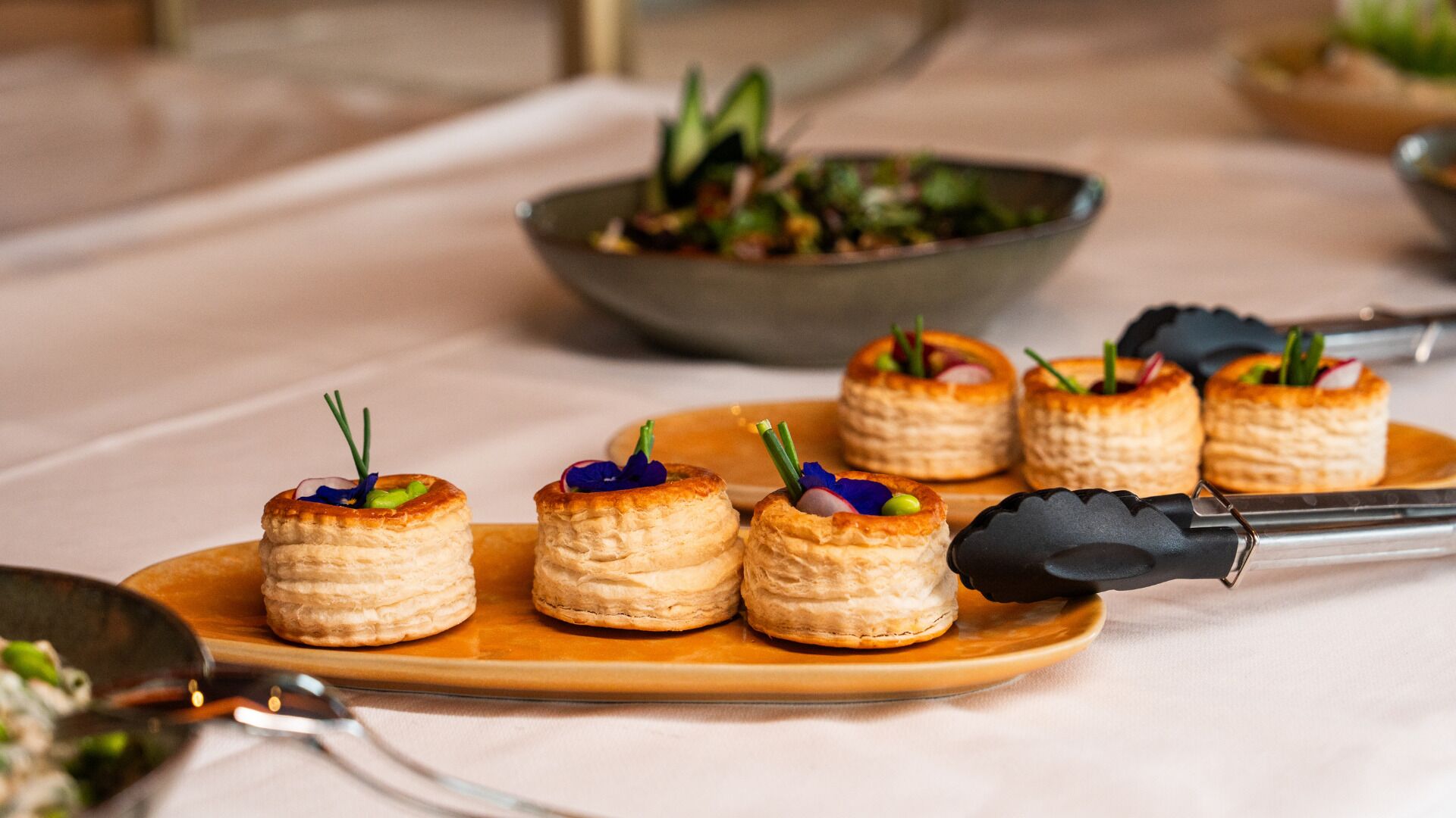A dessert tray with small pastries on a wooden plate, accompanied by a bowl of salad and a pair of tongs.