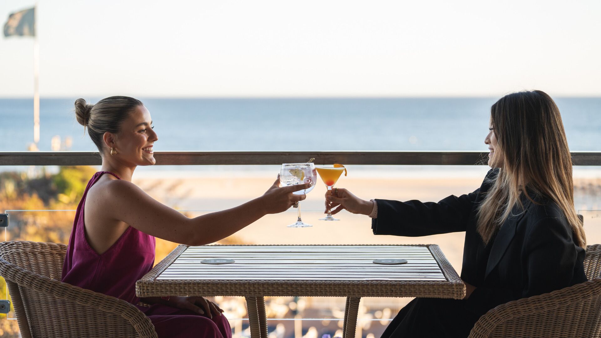 Twee personen proosten aan een tafel met drankjes, met uitzicht op een strand.
