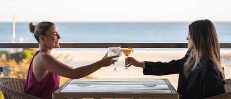 Twee personen proosten aan een tafel met drankjes, met uitzicht op een strand.