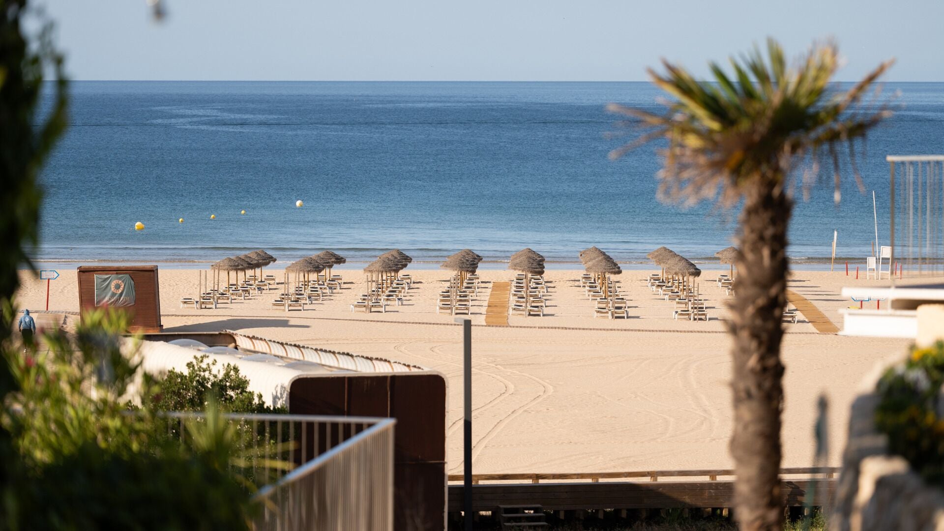 A beach with rows of sun loungers and thatched umbrellas, a wooden deck, and a clear view of the sea.