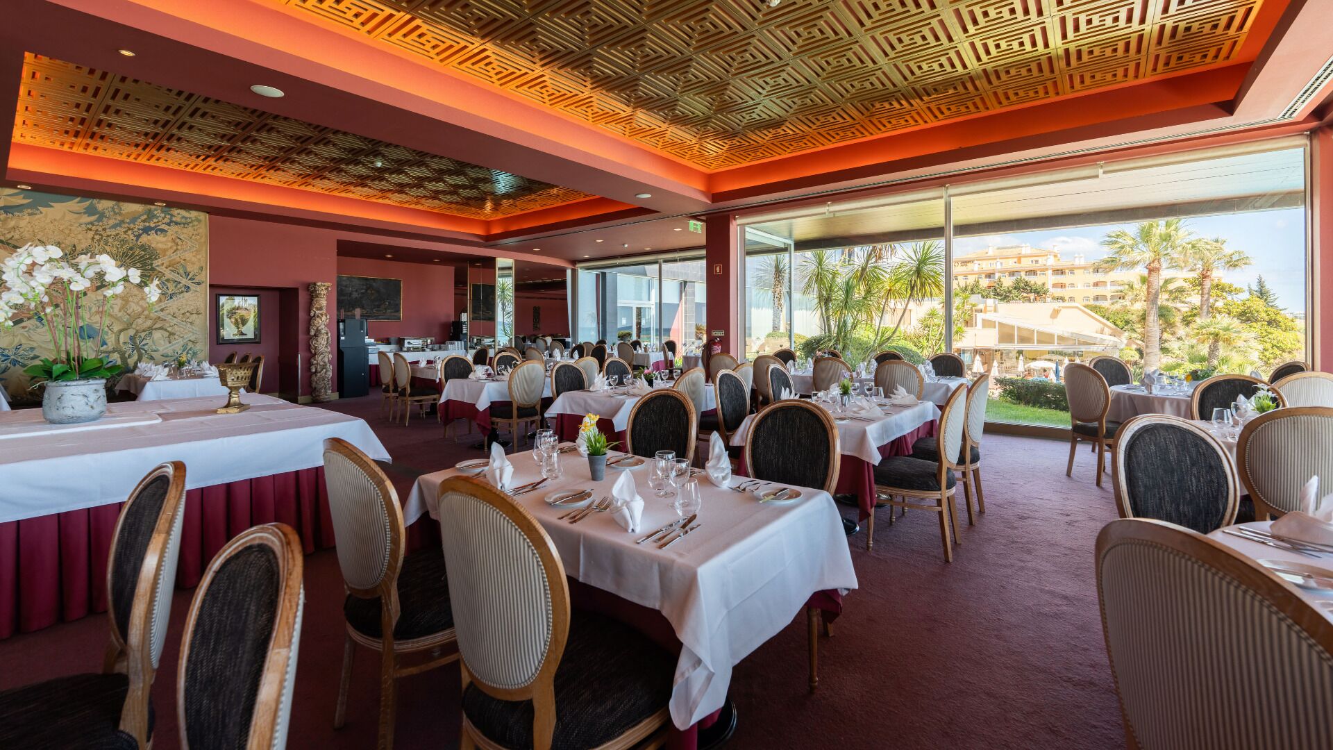 A restaurant with multiple tables set for dining, large windows offering a view of palm trees and buildings, and a decorative ceiling.