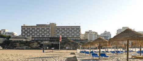 A beach with blue lounge chairs and thatched umbrellas, a wooden walkway, and a large hotel building in the background.
