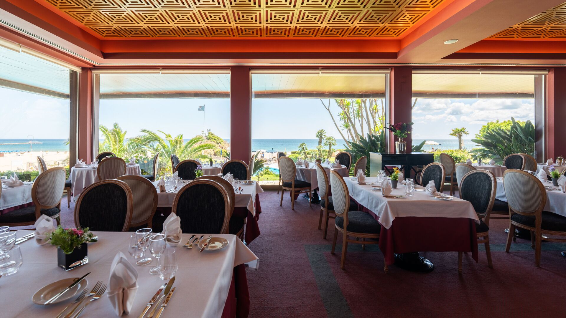 A restaurant with tables set for dining, large windows offering a view of the beach and ocean, and a patterned ceiling.