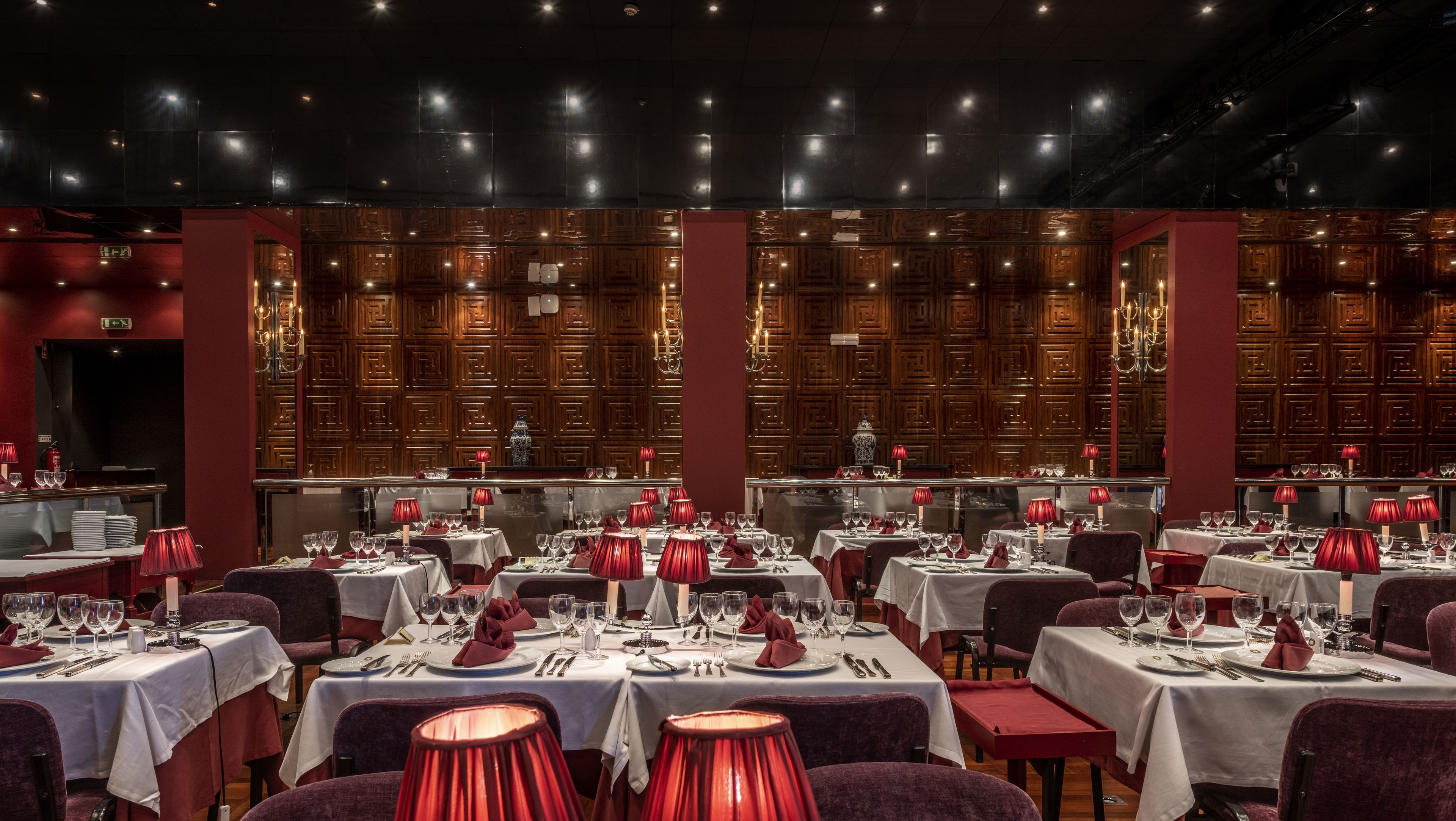 A restaurant with multiple tables set for dining, featuring red tablecloths and red lamps.
