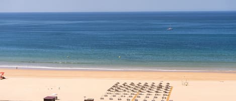 A beach with a row of umbrellas and lounge chairs, a small building, and a boat in the water.