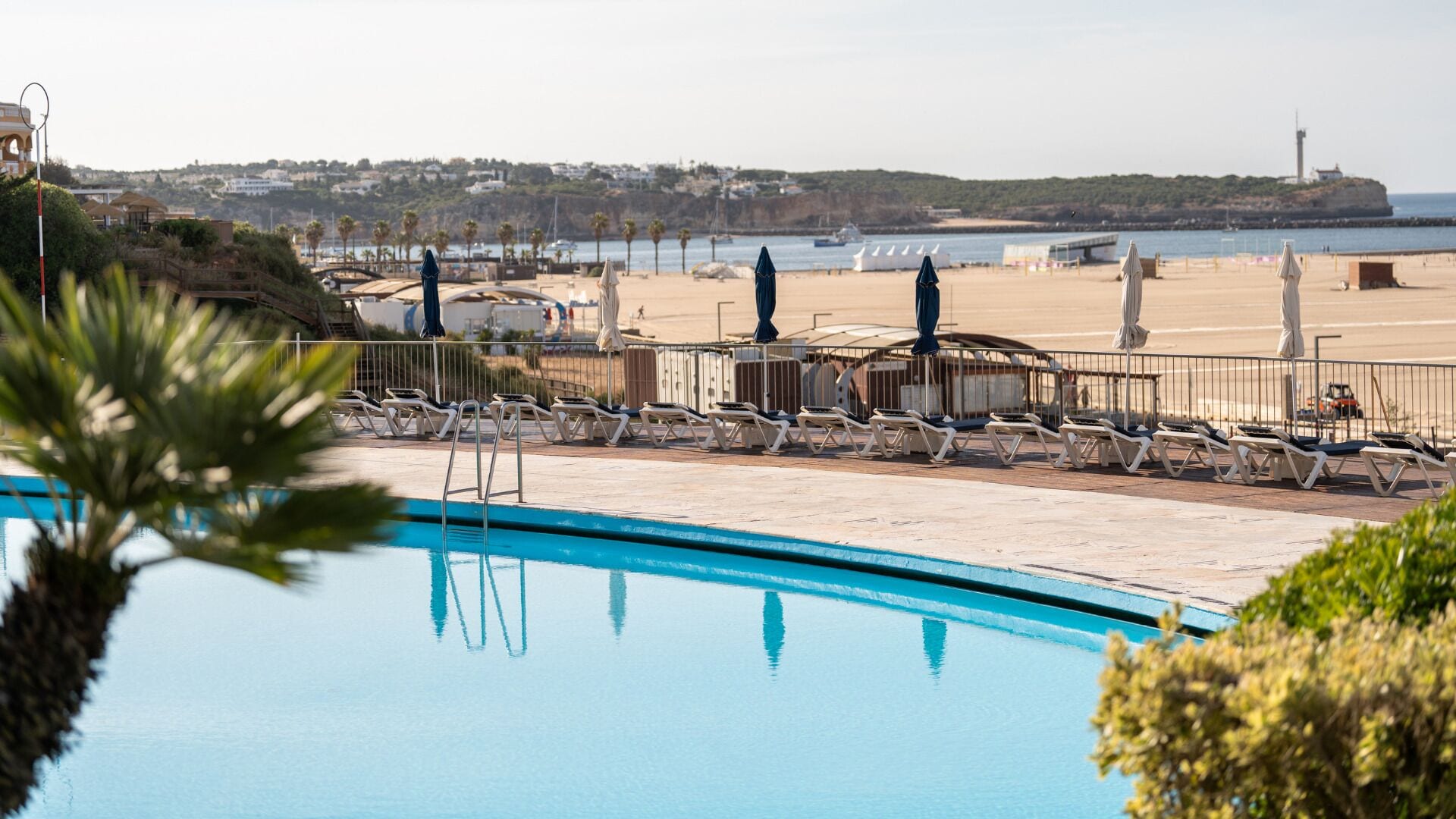 A swimming pool with sun loungers and umbrellas, a beach area with chairs and umbrellas, and a distant view of boats and buildings.