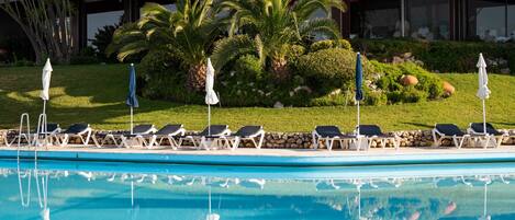 A pool area with lounge chairs and umbrellas, surrounded by greenery and a building in the background.