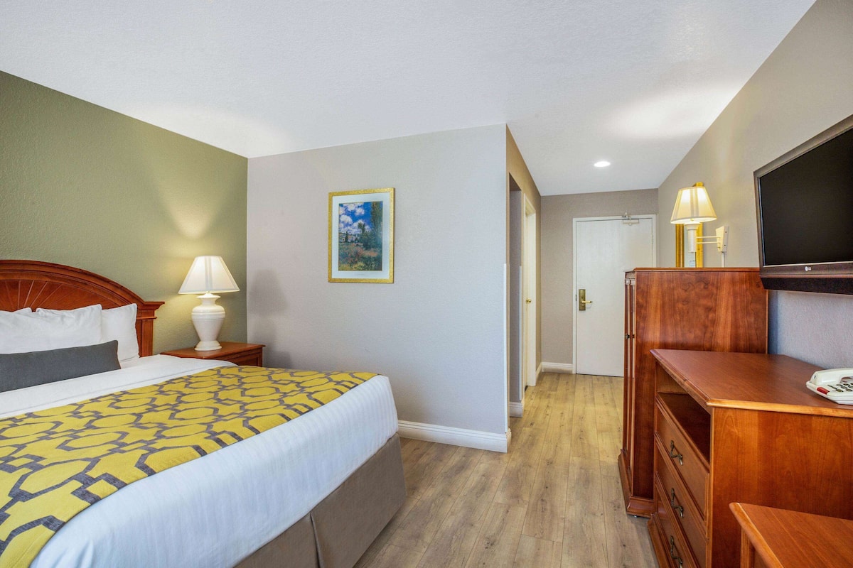 Una habitación de hotel con cama, lámparas de noche, cómoda, televisor y puerta que da a otra estancia.