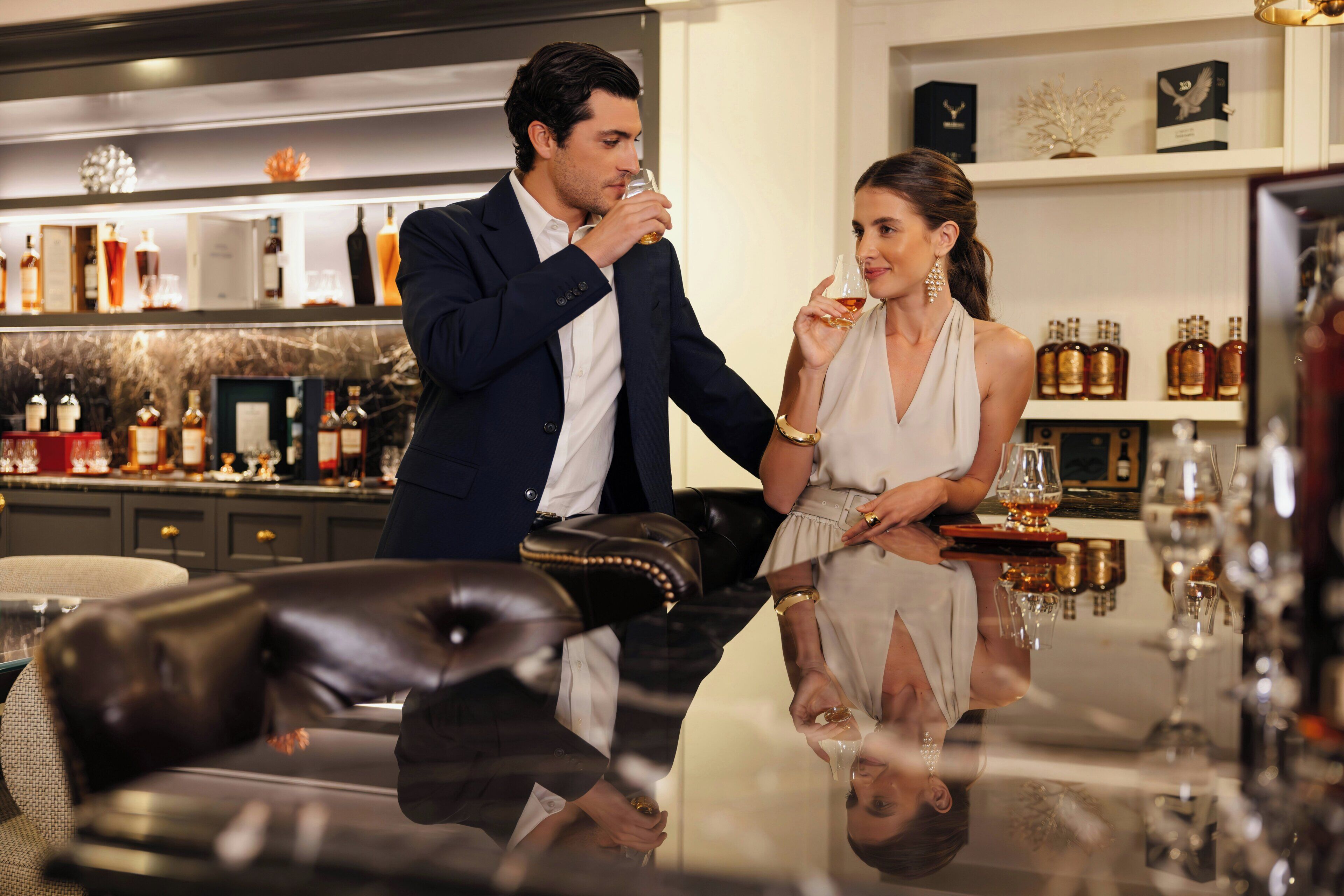 A man and a woman in a bar setting with a glass table, surrounded by various bottles and glasses.