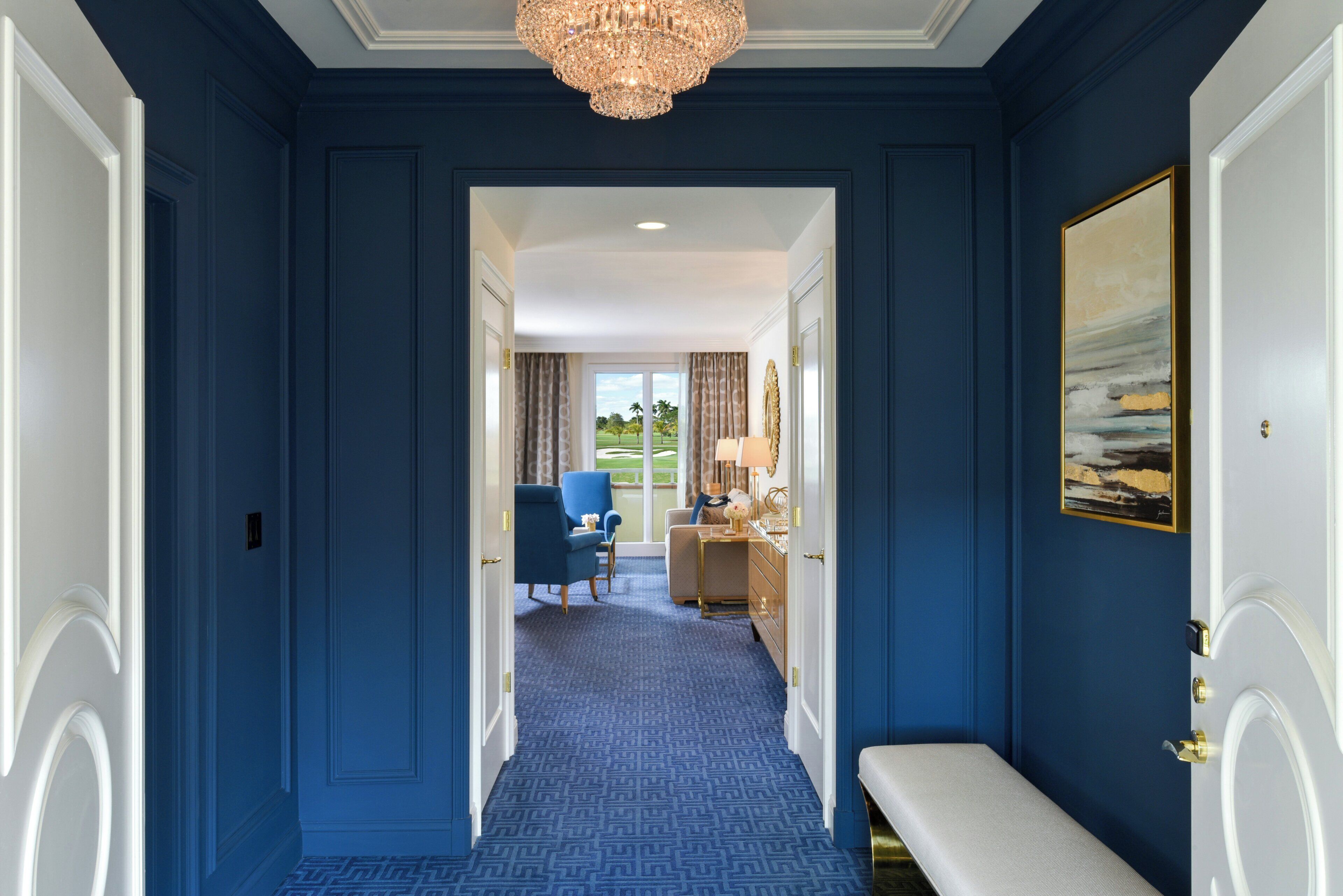 Een gang met blauwe wanden, een kroonluchter, een bankje en uitzicht op een kamer met een bureau en stoel.