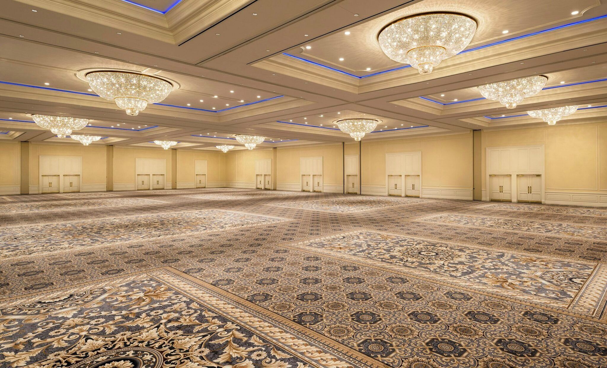 Un amplio salón de conferencias con candelabros ornamentados, alfombra estampada y una disposición simétrica de puertas.