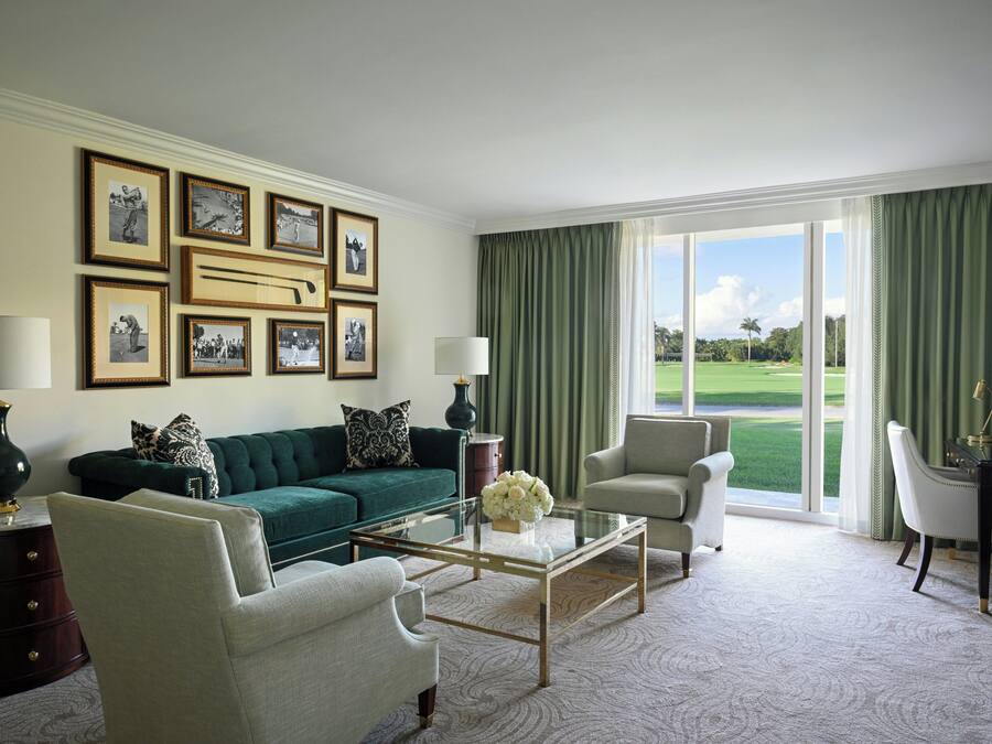 A living room with a green sofa, two armchairs, a glass coffee table, and a view of a golf course through large windows.