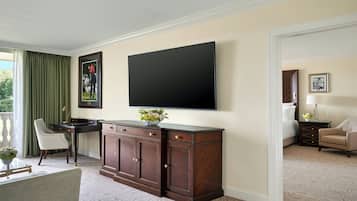 Quarto de hotel com uma televisão de tela grande fixada na parede, um armário de madeira com gavetas, uma mesa, uma cadeira e uma cama com mesa de cabeceira.