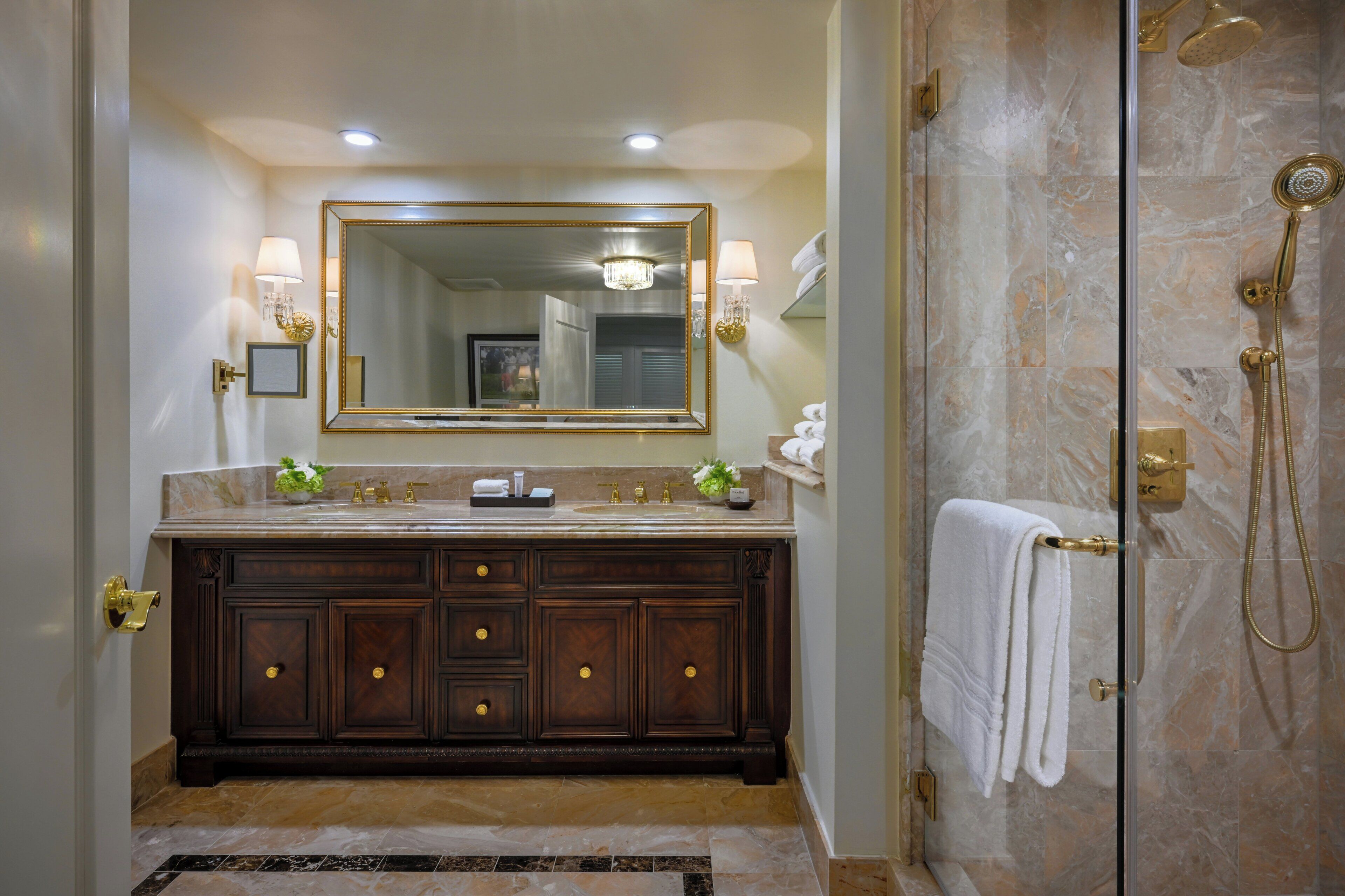 Une salle de bain avec un lavabo en marbre, un grand miroir, une douche dotée d’un pommeau de douche doré et un porte-serviettes.