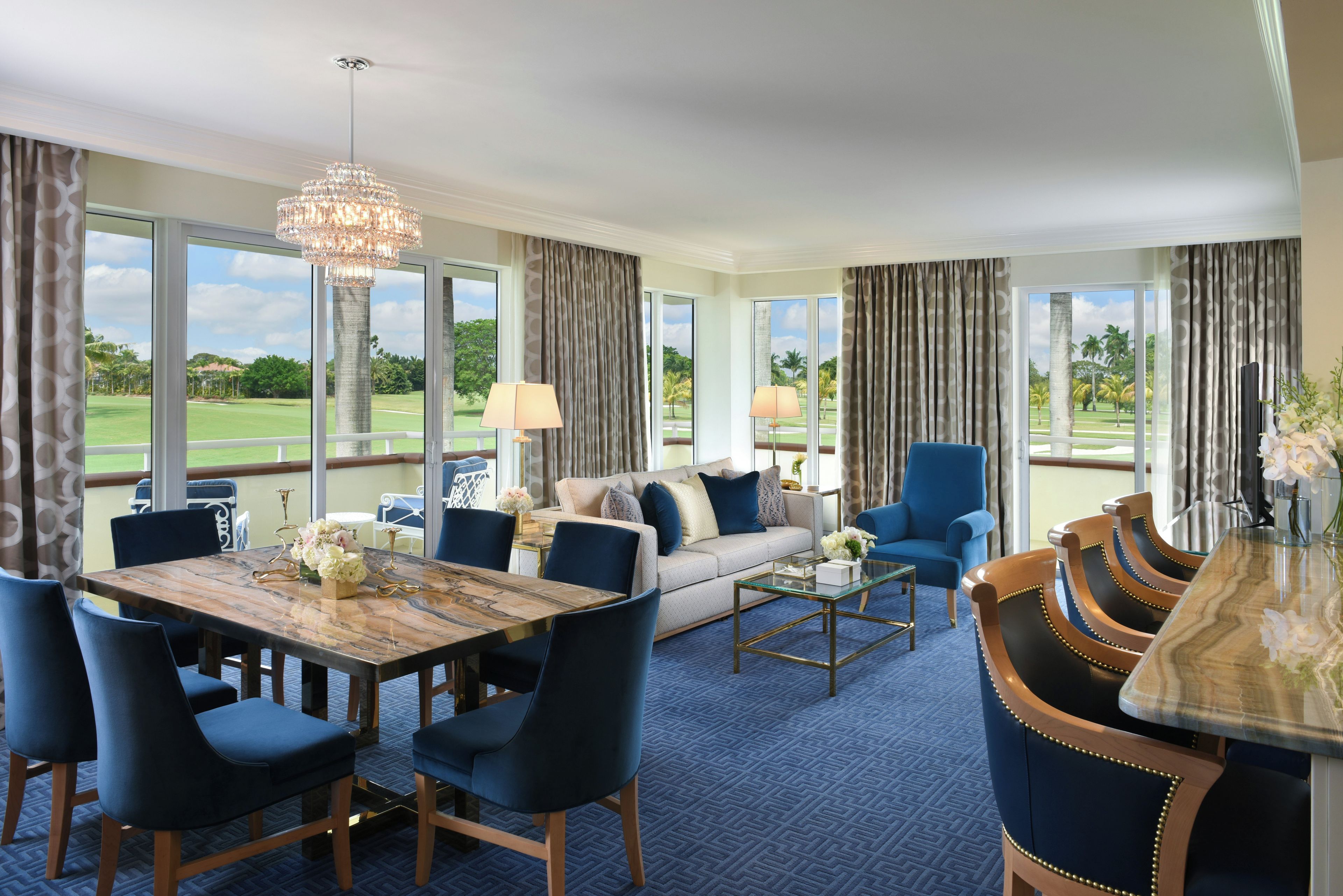 Une chambre d’hôtel comprenant un coin repas, un ensemble de canapés, un lustre et de grandes fenêtres donnant sur un terrain de golf.
