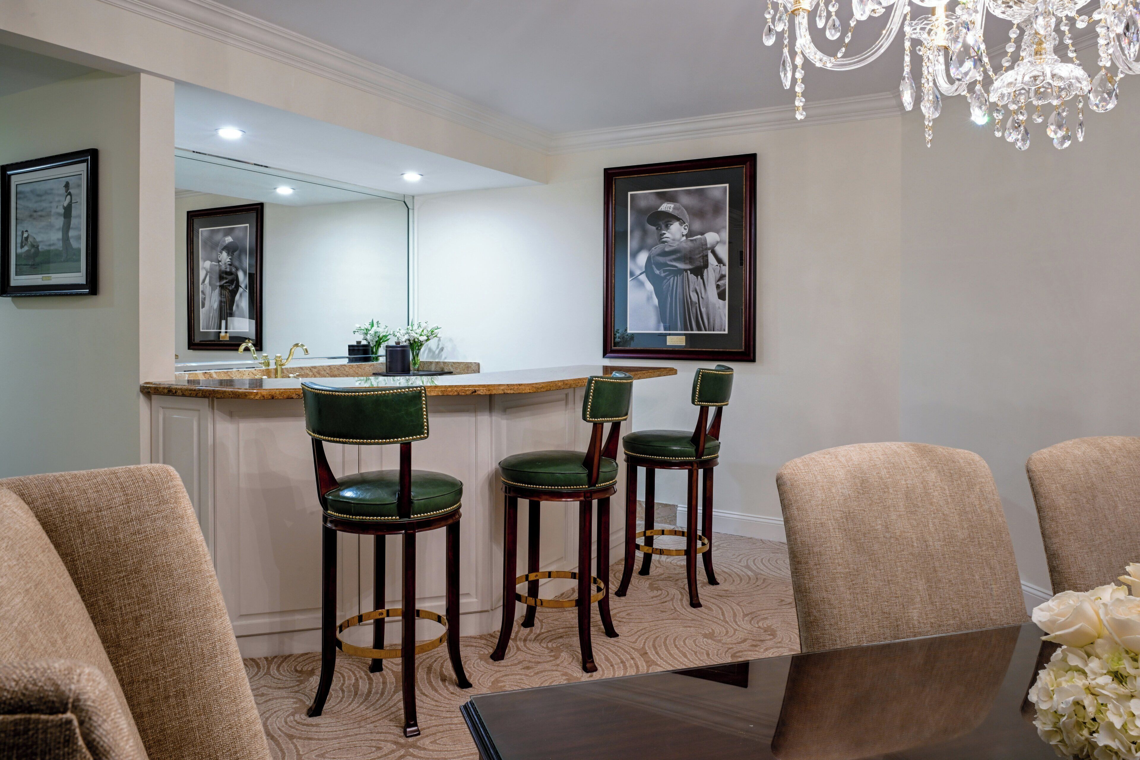 A dining area with a bar, green upholstered chairs, a wooden table, and framed artwork on the walls.