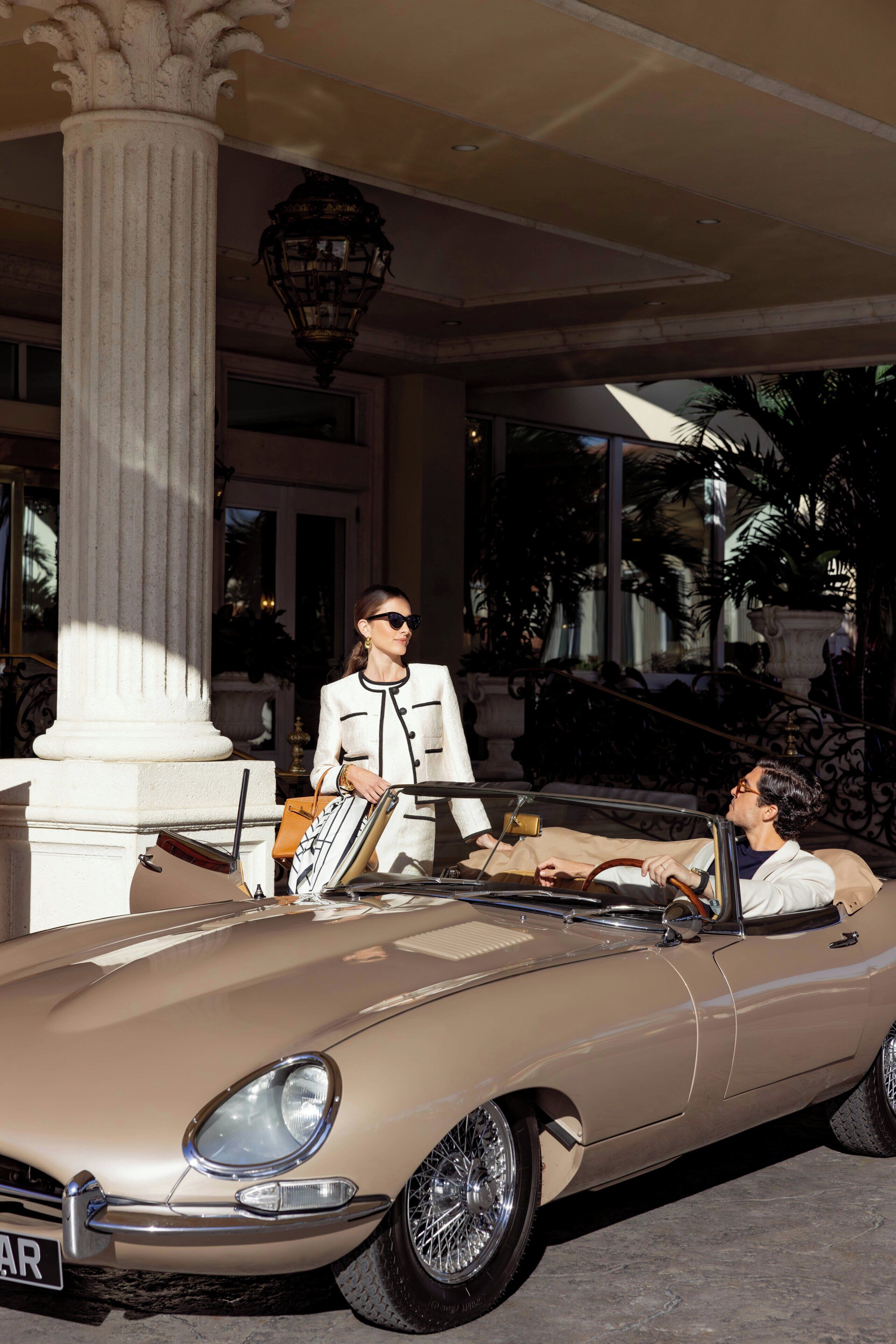 A classic convertible car parked in front of a building with a large column and a decorative lamp.
