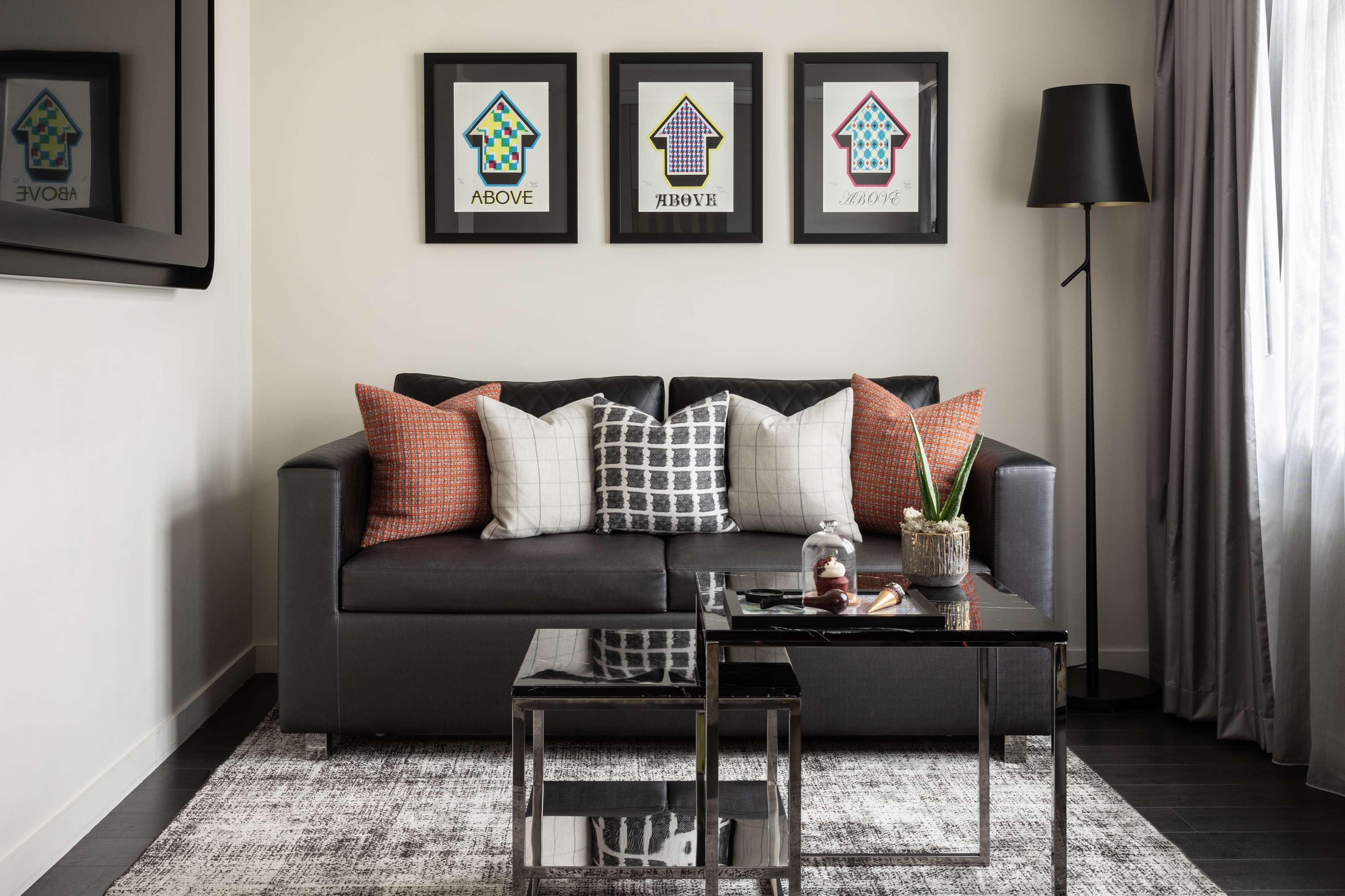 A modern living room with a dark grey sofa, patterned cushions, a glass coffee table, and framed artwork on the wall.