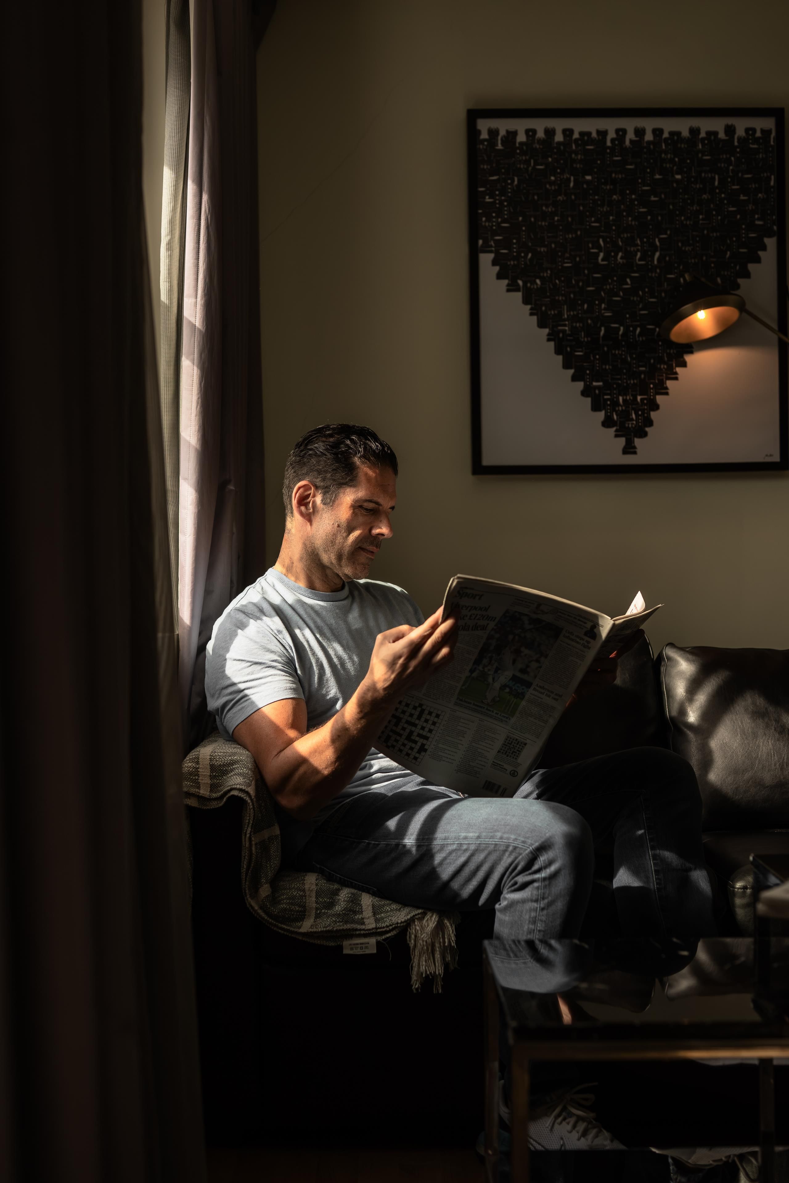 Une personne est assise sur une chaise, lisant un journal dans une pièce éclairée par une lampe, et un tableau encadré est accroché au mur.